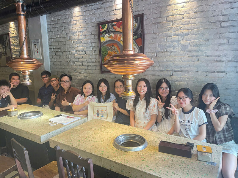 Thirteen members of the unit seated together along one side of a long table in a restaurant, several holding up a tote bag printed with a cartoon design.
