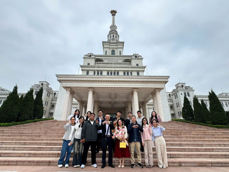 Fifteen members of the unit standing in two rows on the steps of a large white colonnaded building, one of them holding a bouquet of pink and white flowers.
